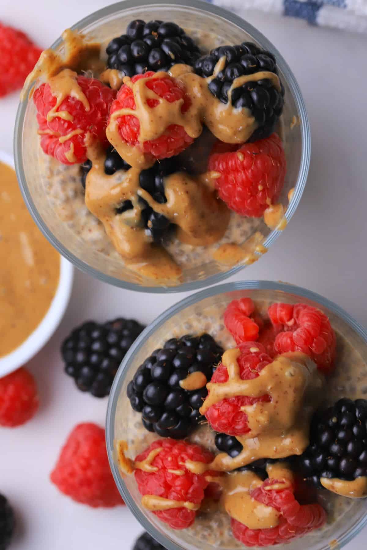 Two cups with low carb chia almond butter pudding topped with berries and almond butter with almond butter and berries on the side.