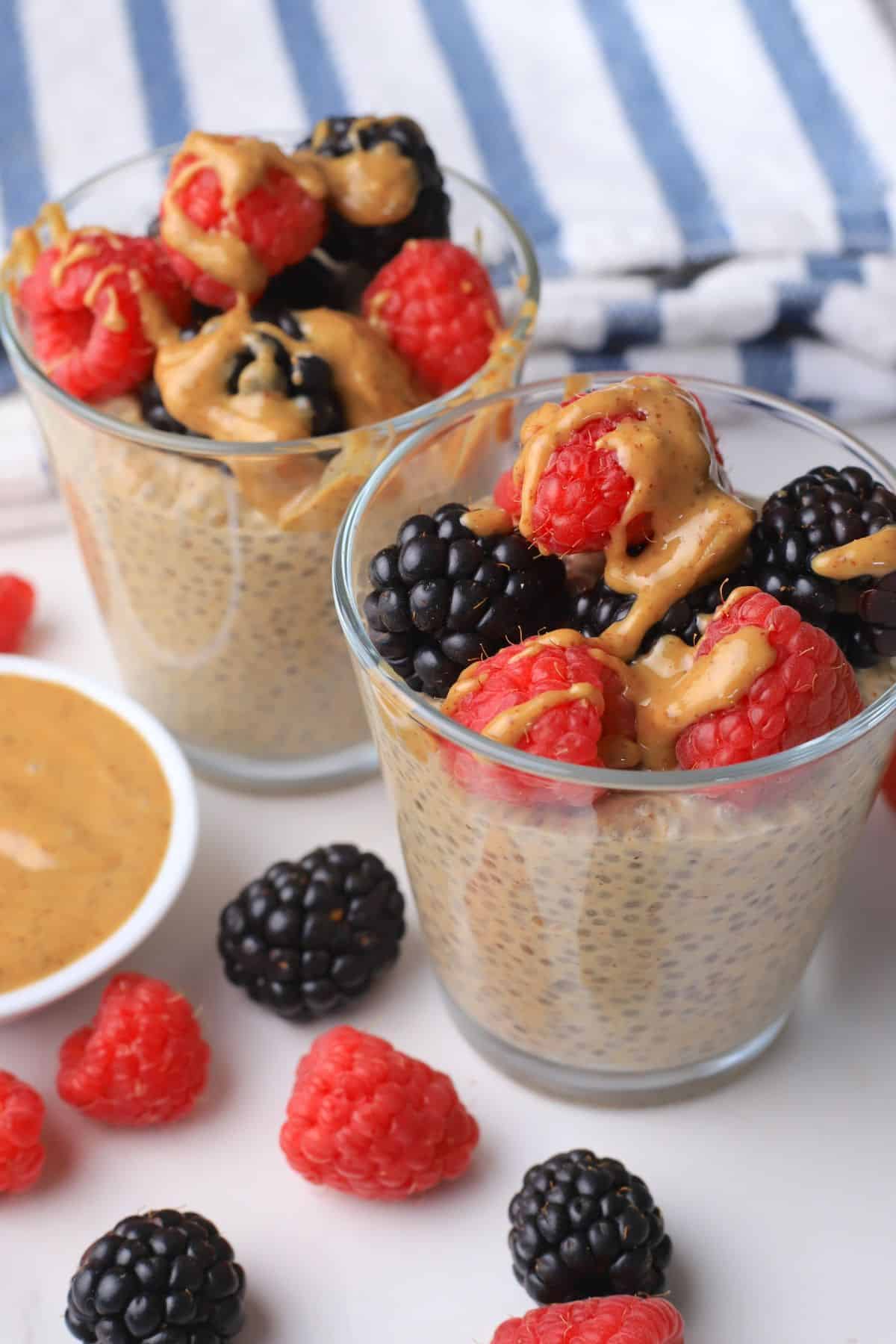 Two cups with low carb chia almond butter pudding topped with berries and almond butter with almond butter and berries on the side.