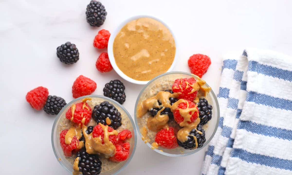 Two cups with low carb chia almond butter pudding topped with berries and almond butter with almond butter and berries on the side.
