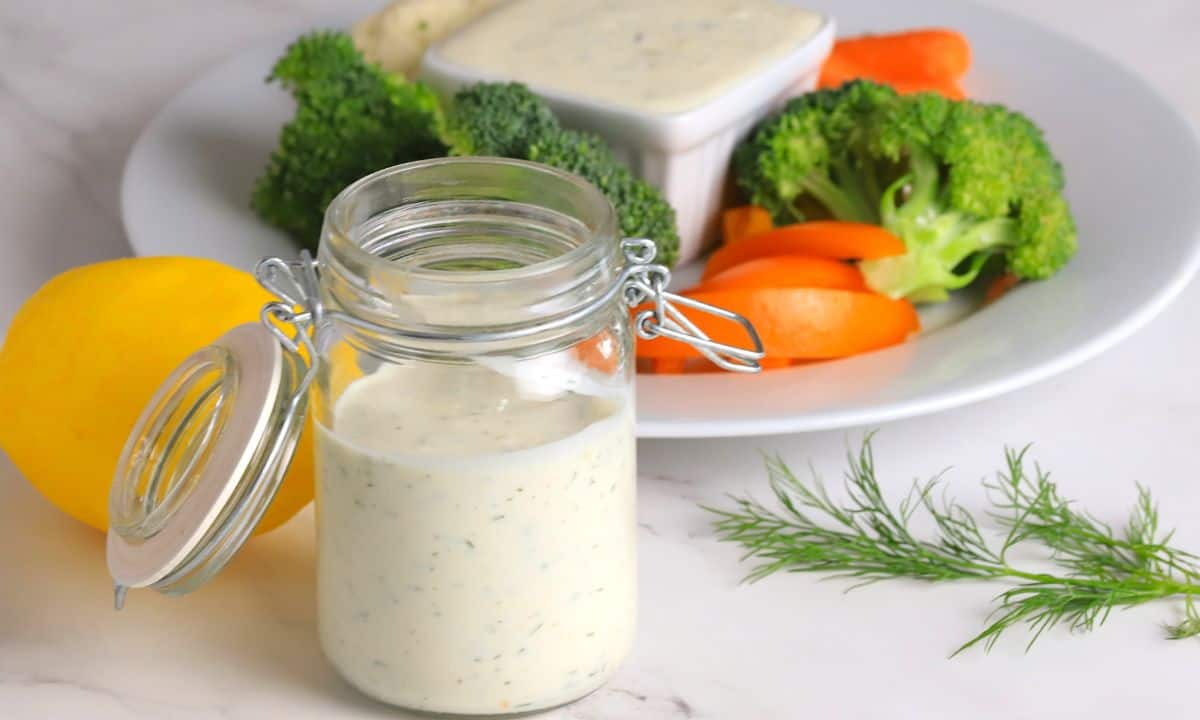 Wide shot of greek yogurt protein ranch in a mason jar next to a plate of vegetables with a ranch dip.