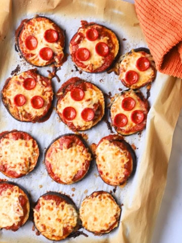 Low carb eggplant pizzas on a parchment lined baking sheet with a an orang dish towel.