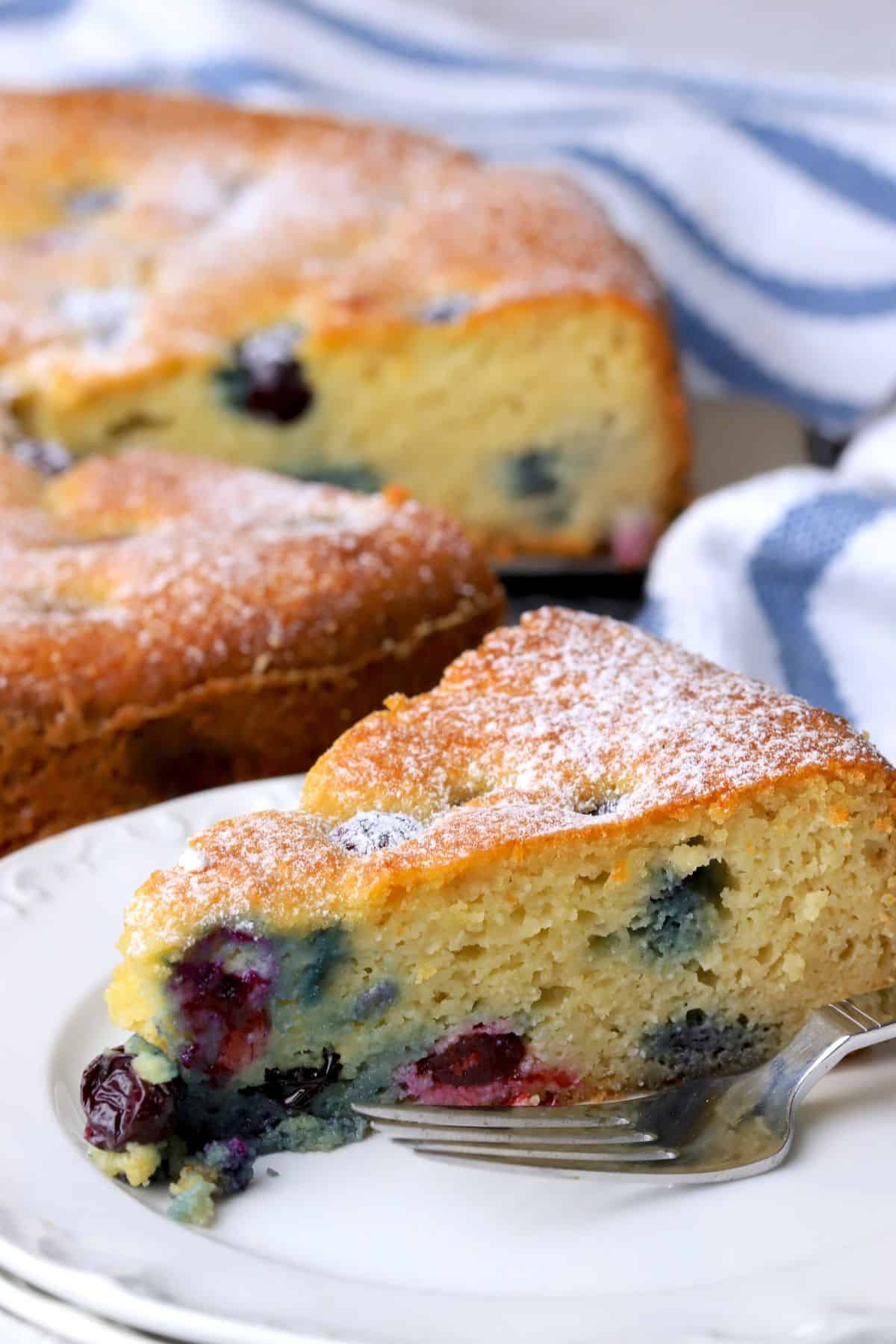 A slice of the low carb ricotta blueberry cake on a plate with a fork, next to the cake.