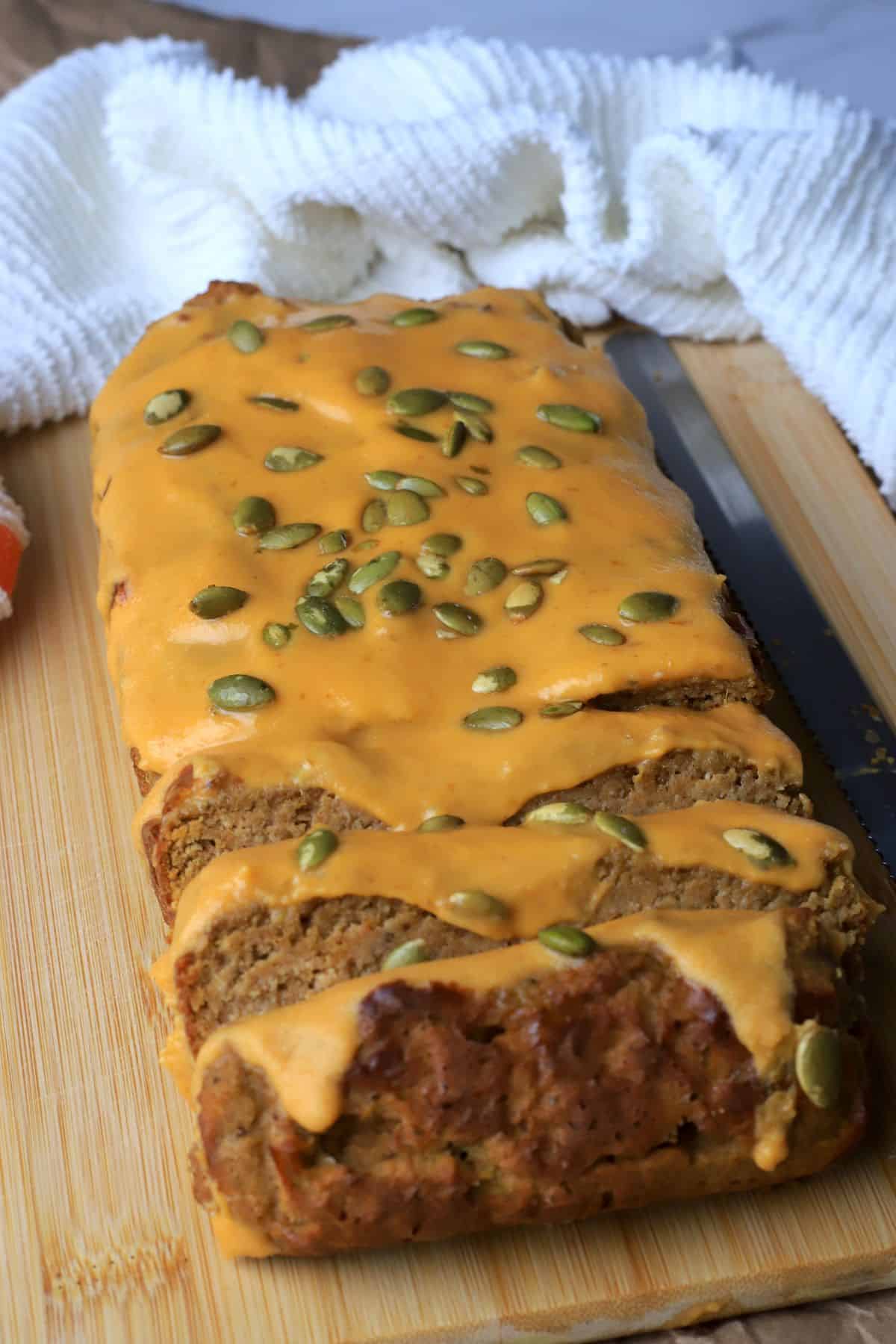 High protein pumpkin spice loaf with a glaze and pumpkin seed topping on a cutting board with a kitchen towel.