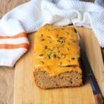 High protein pumpkin spice loaf with a glaze and pumpkin seed topping on a cutting board with a knife.