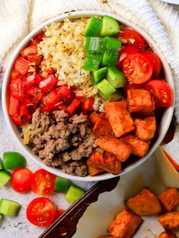 Sweet potato protein bowl next to a kitchen towel, chopped cucumbers, tomatoes and sweet potatoes on baking tray.