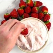 High protein strawberry cheesecake fluff in a bowl, dipping a strawberry and more strawberries surrounding the bowl.