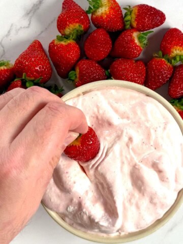 High protein strawberry cheesecake fluff in a bowl, dipping a strawberry and more strawberries surrounding the bowl.