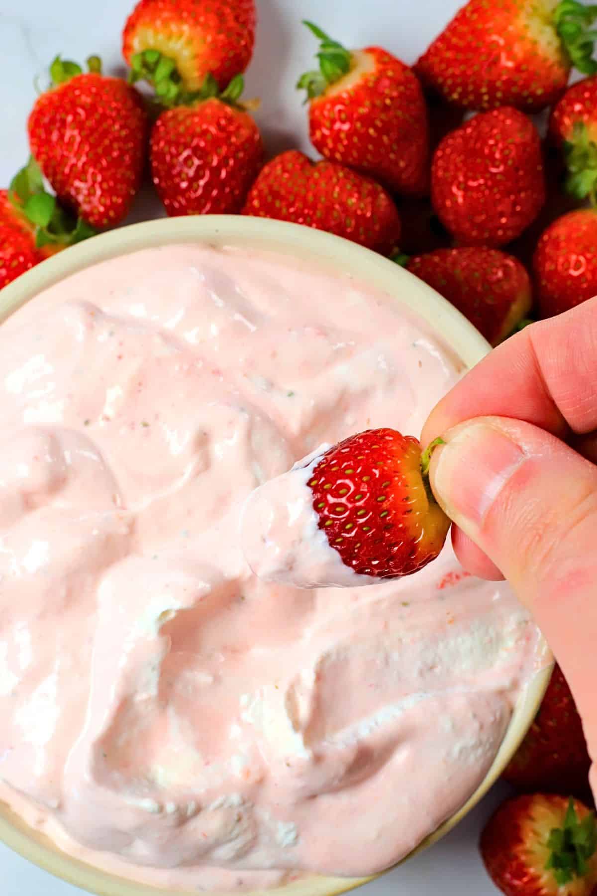 High protein strawberry cheesecake fluff in a bowl, dipping a strawberry and more strawberries surrounding the bowl.