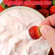 High protein strawberry cheesecake fluff in a bowl, dipping a strawberry and more strawberries surrounding the bowl.