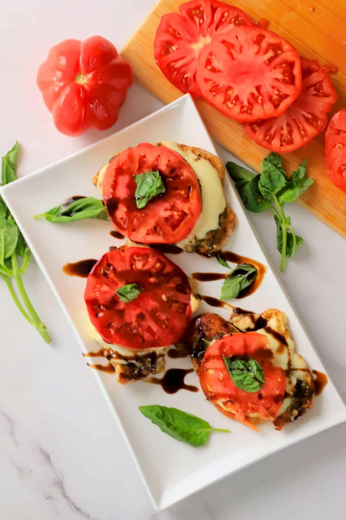 Low carb pesto caprese chicken on a platter next to sliced tomatoes on a cutting board.