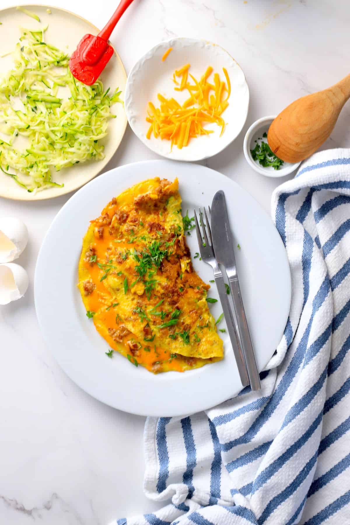 Low carb zucchini sausage omelette on a plate next to a plate with shredded zucchini and another with shredded cheddar.