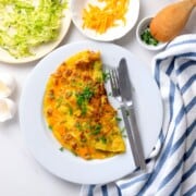 Low carb zucchini sausage omelette on a plate next to a plate with shredded zucchini and another with shredded cheddar.