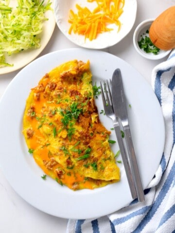 Low carb zucchini sausage omelette on a plate next to a plate with shredded zucchini and another with shredded cheddar.
