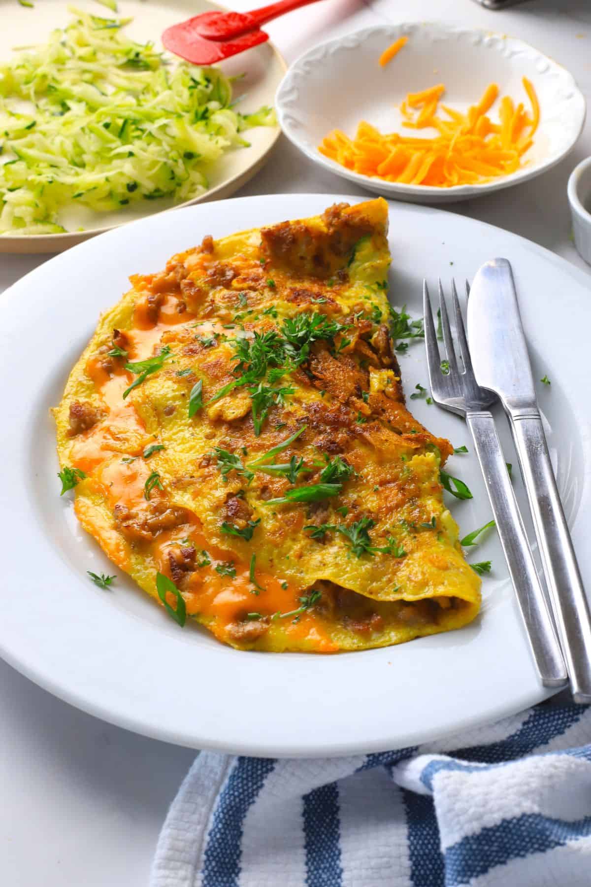 Low carb zucchini sausage omelette on a plate next to a plate with shredded zucchini and another with shredded cheddar.