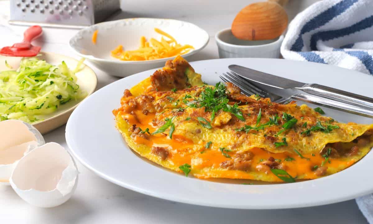 Low carb zucchini sausage omelette on a plate next to a plate with shredded zucchini and another with shredded cheddar.