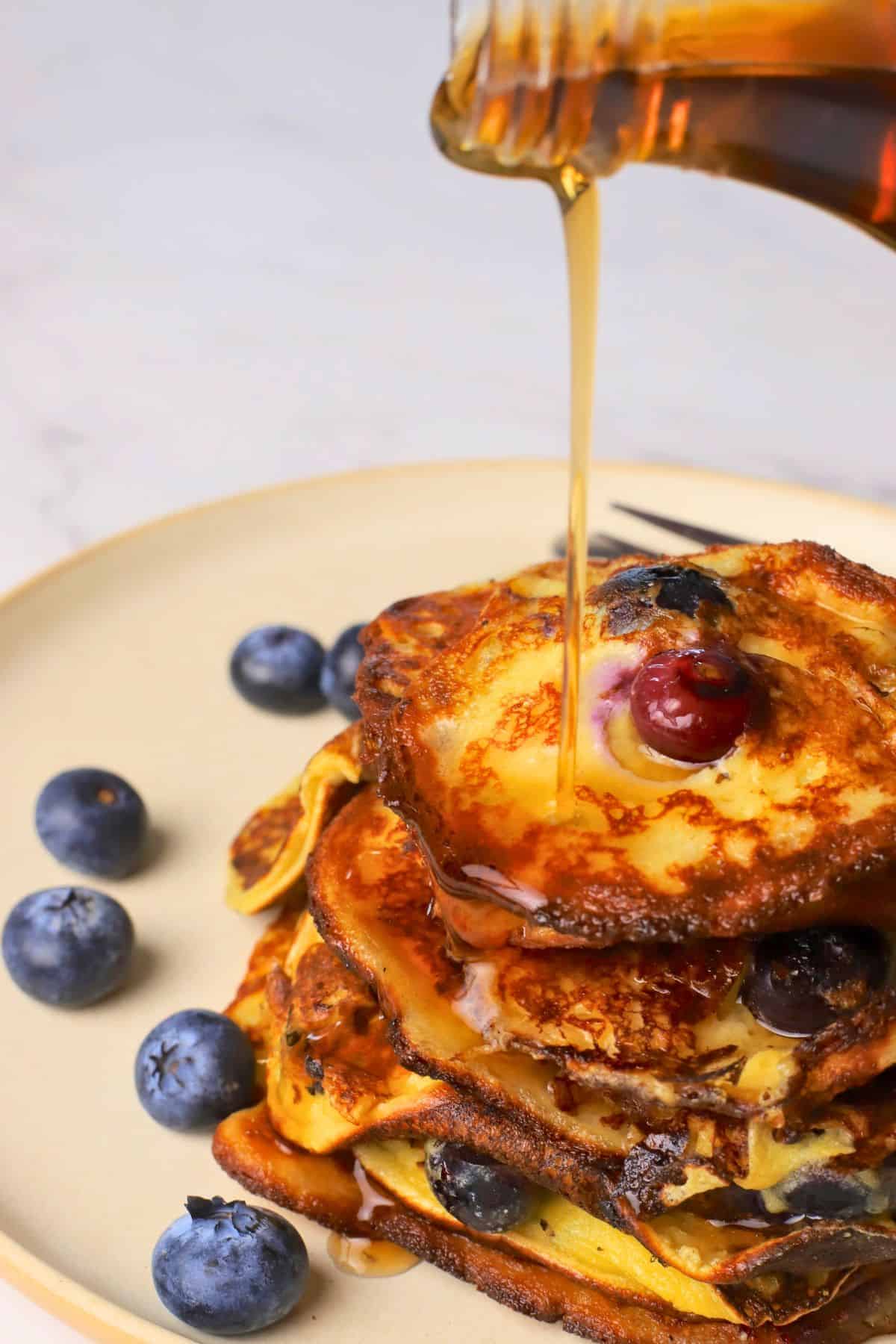 Pouring syrup over blueberry cottage cheese pancakes served on a plate with fresh blueberries.