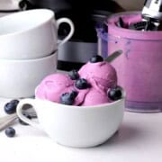 Low carb blueberry cottage cheese ice cream scoops in a bowl topped with fresh blueberries. The ninja cream pint and bowls in the background.