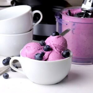 Low carb blueberry cottage cheese ice cream scoops in a bowl topped with fresh blueberries. The ninja cream pint and bowls in the background.