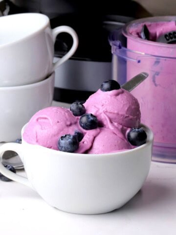 Low carb blueberry cottage cheese ice cream scoops in a bowl topped with fresh blueberries. The ninja cream pint and bowls in the background.