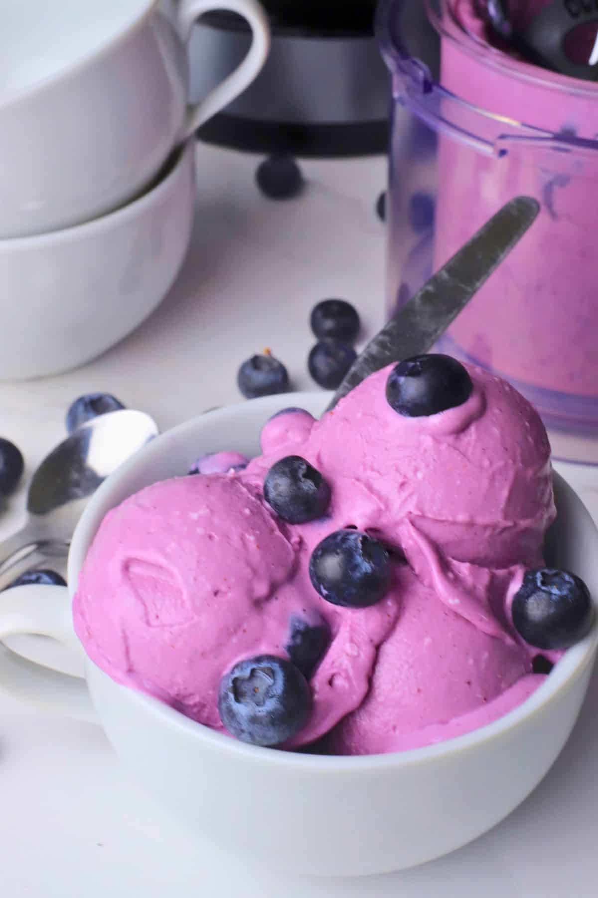 Low carb blueberry cottage cheese ice cream scoops in a bowl topped with fresh blueberries. The ninja cream pint and bowls in the background.