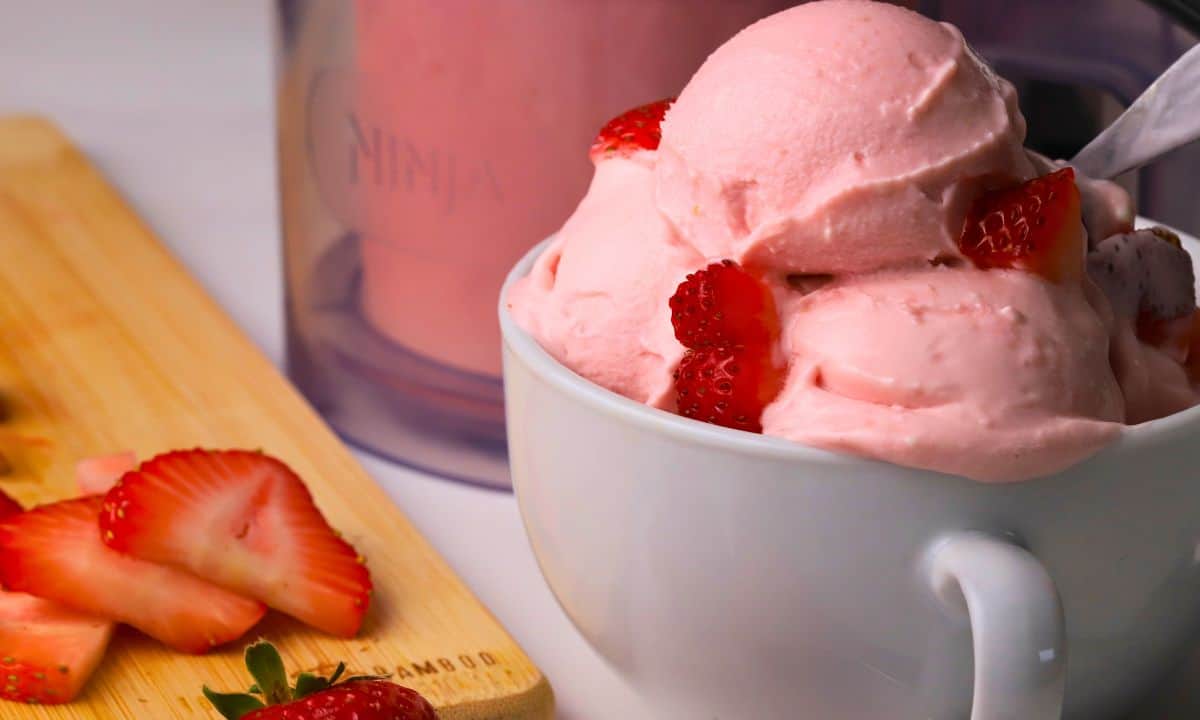 Low carb strawberry cottage cheese ice cream in a bowl with strawberries and the ninja creami pint next to the bowl.