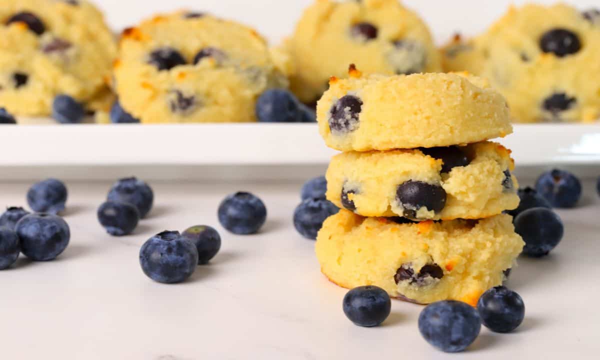 Three low carb blueberry cottage cheese cookies stacked on top of each other next to a plate of the same.
