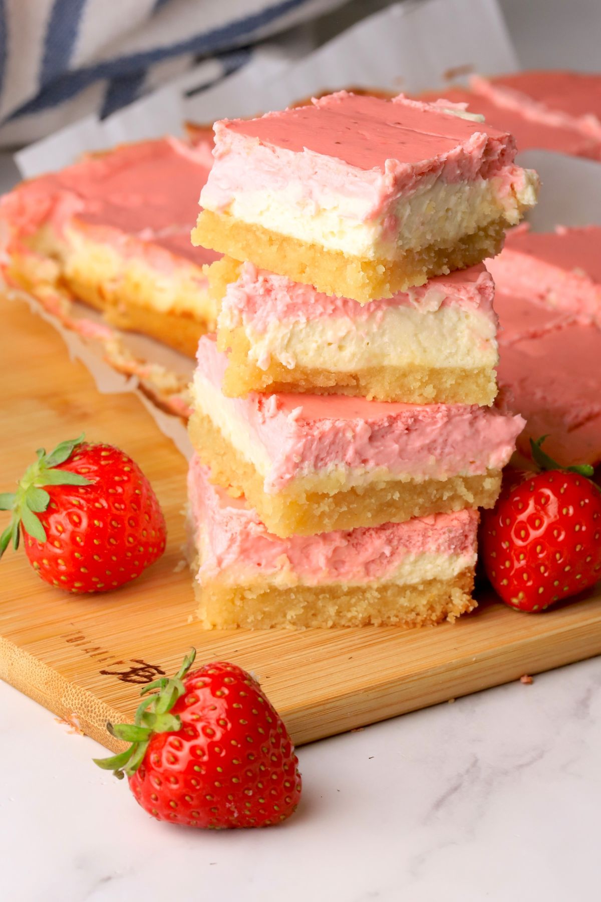 Low carb strawberry cheesecake bars stacked on top of each other next to more sliced bars on a cutting board with fresh strawberries.