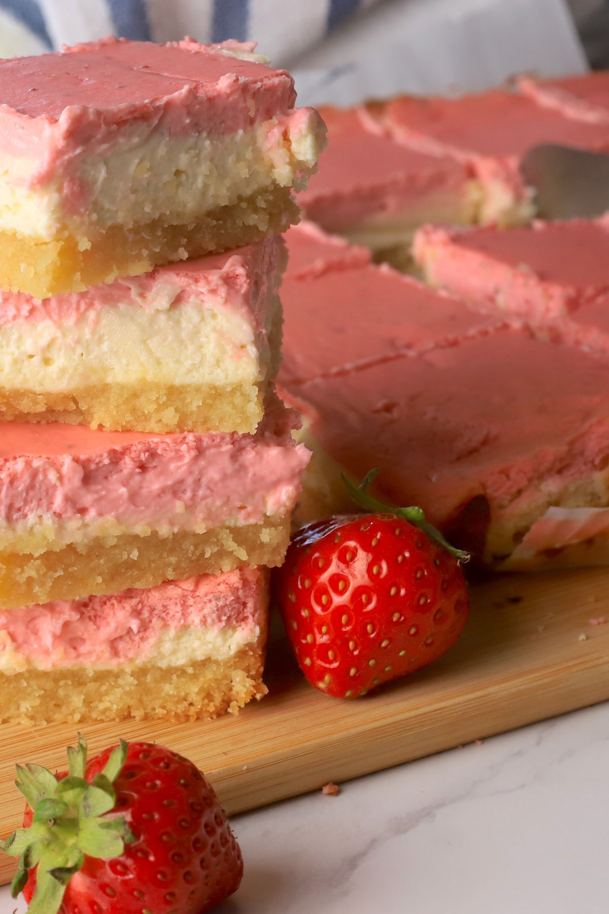 Low carb strawberry cheesecake bars stacked on top of each other next to more sliced bars on a cutting board with fresh strawberries.