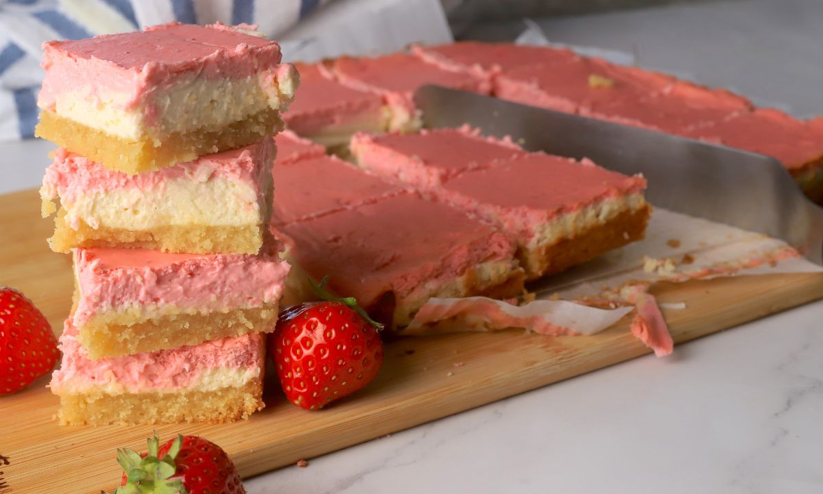 Low carb strawberry cheesecake bars stacked on top of each other next to more sliced bars on a cutting board with fresh strawberries.