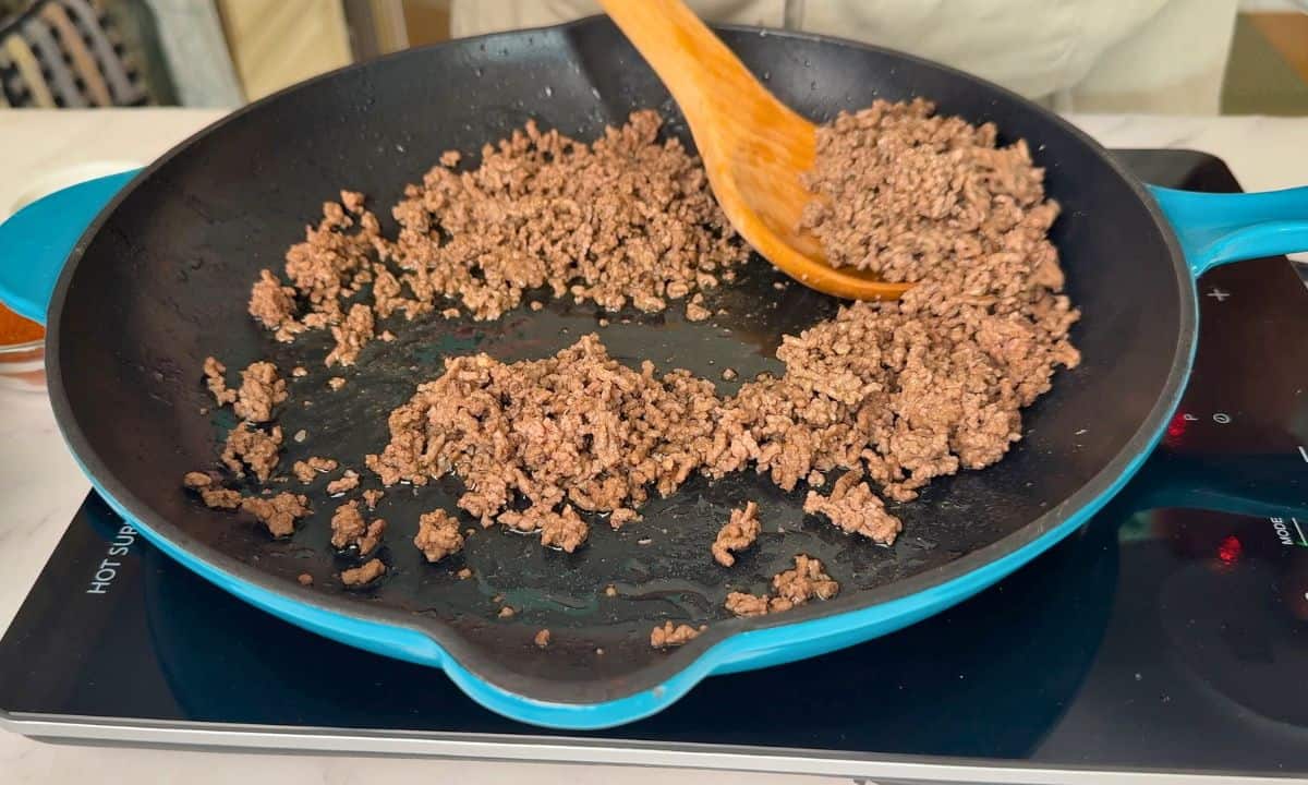 Removing the browned ground meat from the skillet with a wooden spoon.