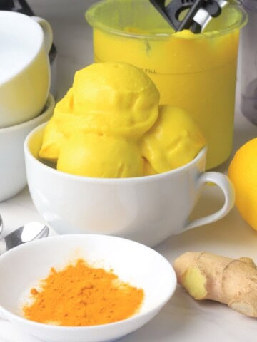 Low carb lemon turmeric ice cream in a bowl next to a lemon, turmeric spices and ginger.