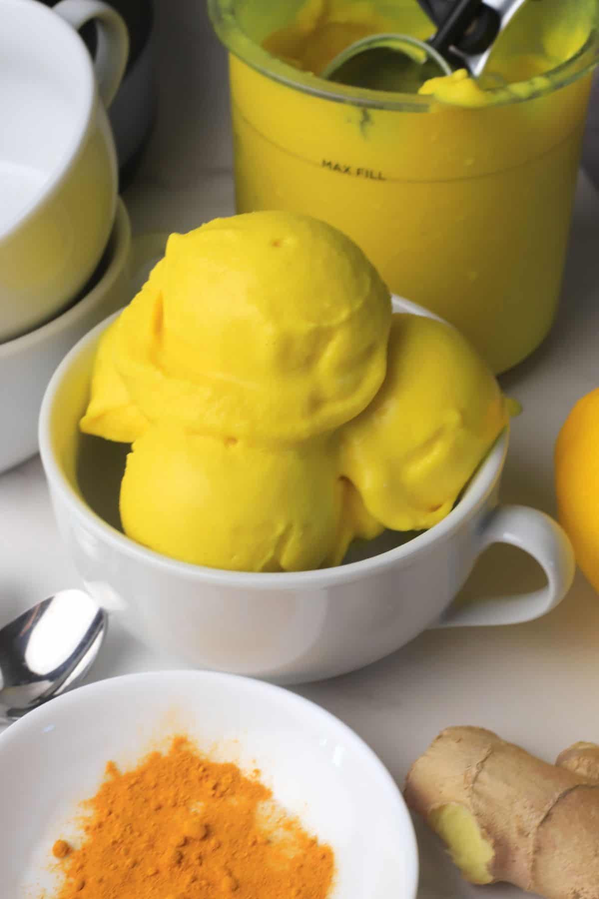 Low carb lemon turmeric ice cream in a bowl next to a lemon, turmeric spices and ginger.
