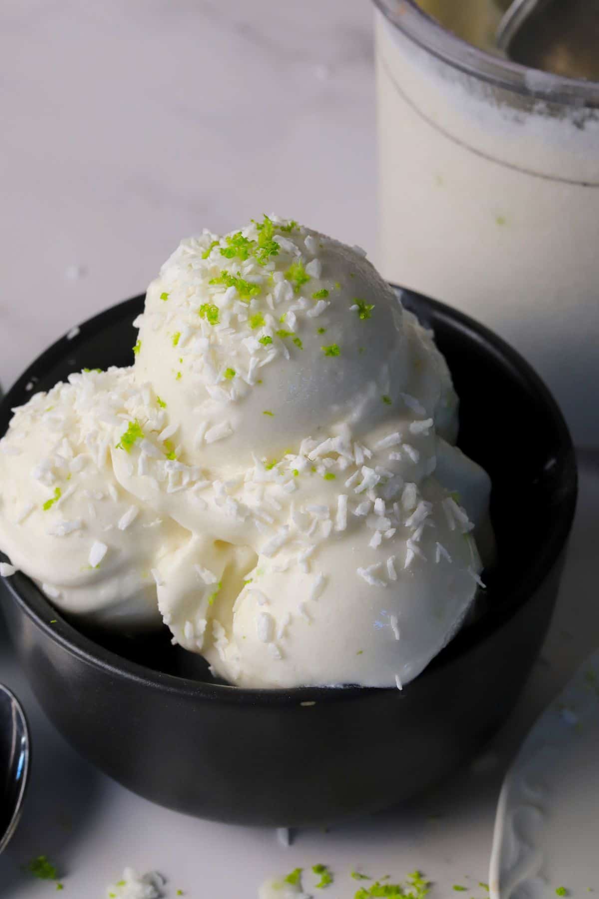 Scoops of low carb coconut lime ice cream in a black bowl with the ninja creami pint in the background.