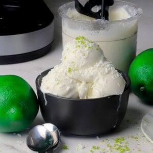 Scoops of low carb coconut lime ice cream in a black bowl with the ninja creami pint in the background with some limes.