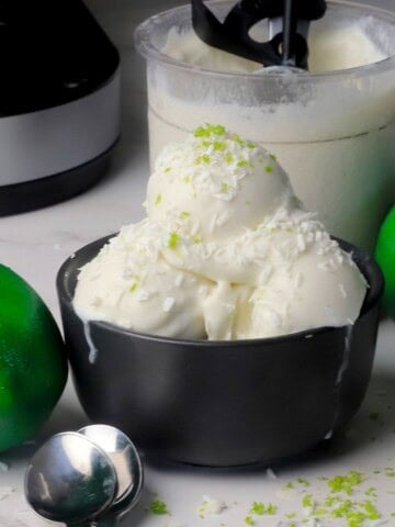 Scoops of low carb coconut lime ice cream in a black bowl with the ninja creami pint in the background with some limes.