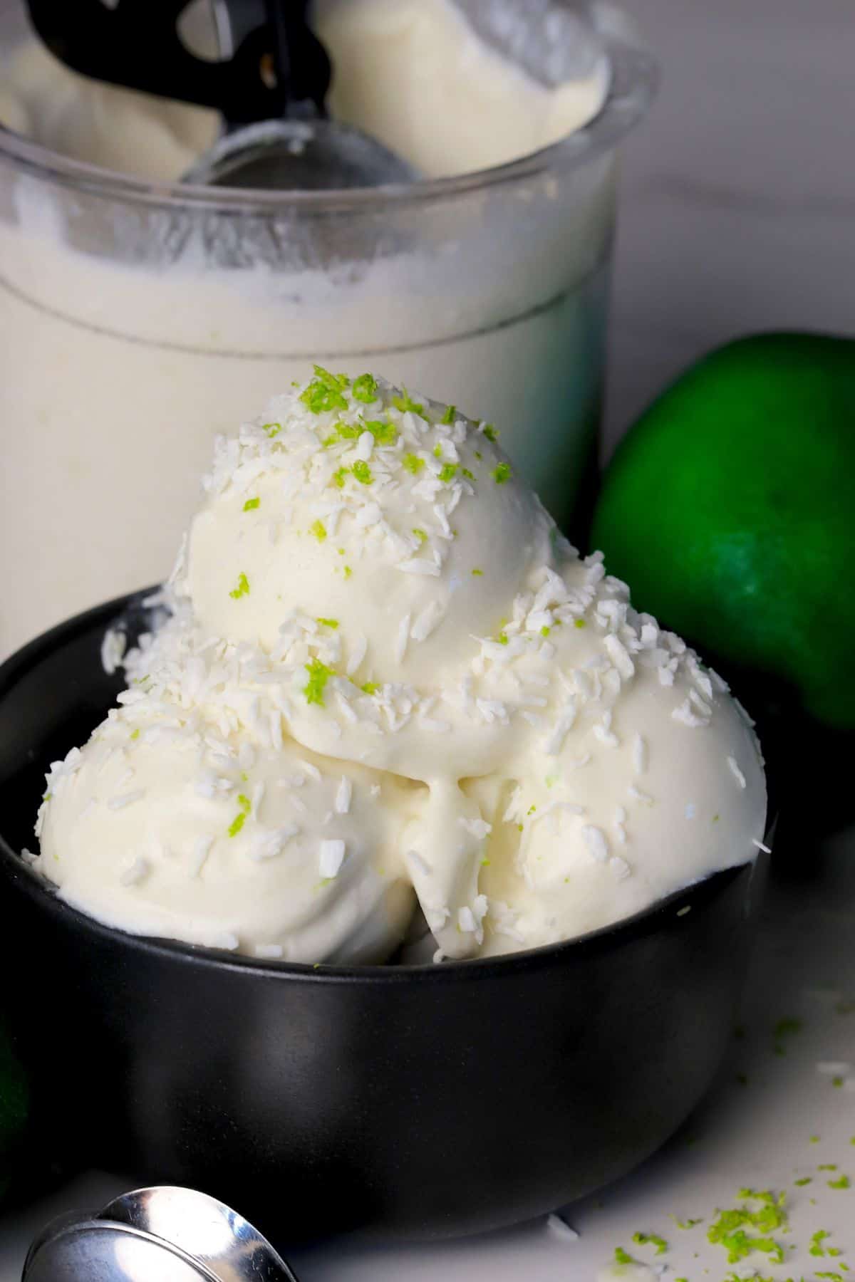 Scoops of low carb coconut lime ice cream in a black bowl with the ninja creami pint in the background with some limes.