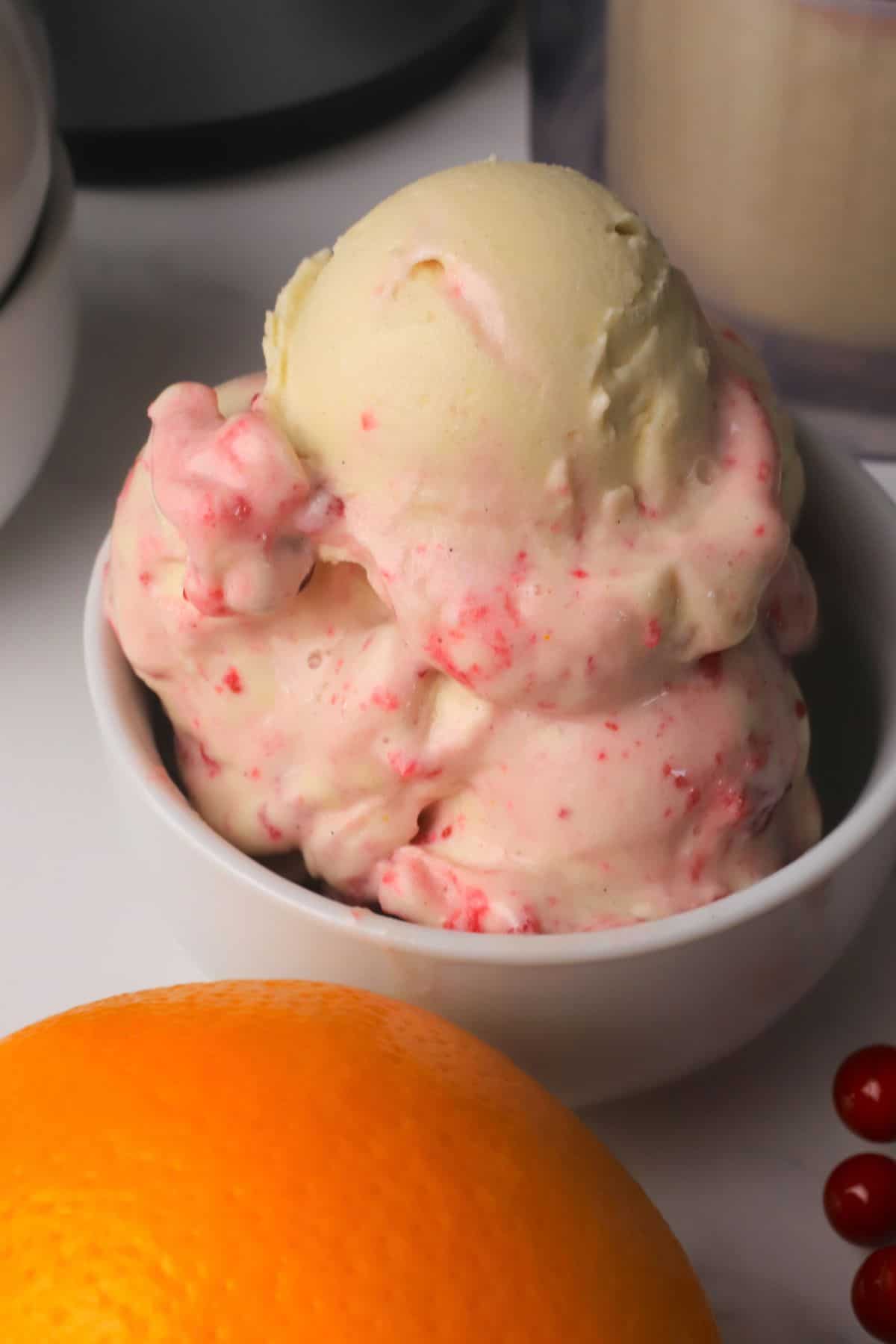 Scoops of low carb cranberry orange spice ice cream in a bowl next to the ninja creami pint and an orange.