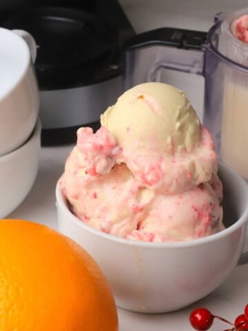 Scoops of low carb cranberry orange spice ice cream in a bowl next to the ninja creami pint and an orange.