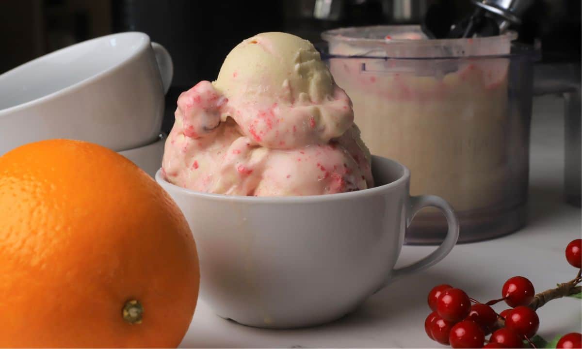 Scoops of low carb cranberry orange spice ice cream in a bowl next to the ninja creami pint and an orange.